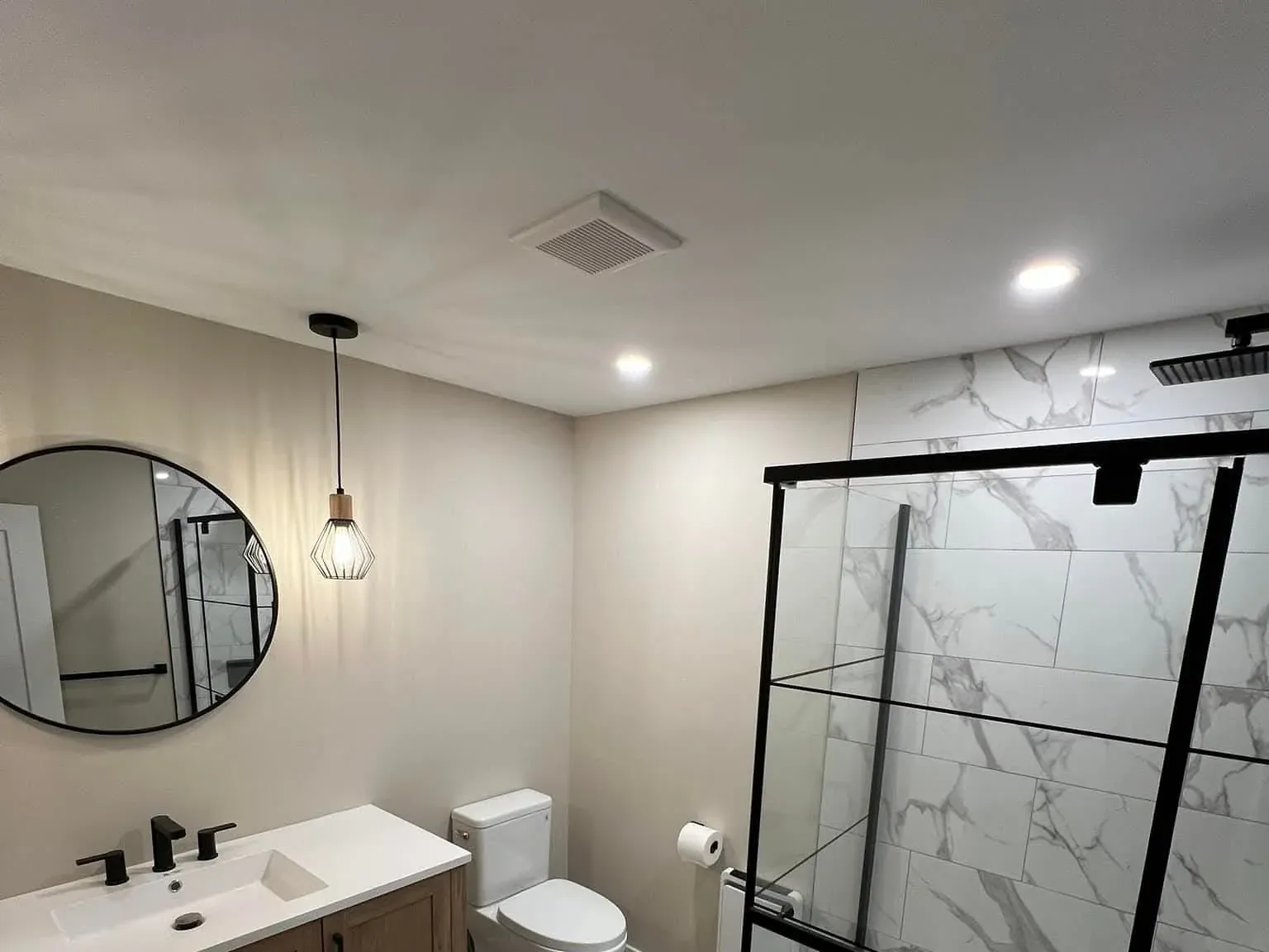 Renovated bathroom with a black-framed glass shower, marble-look tile, a wood vanity, and a round mirror